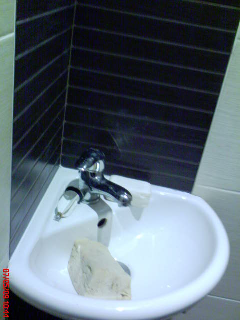 Narożna umywalka z chromowaną baterią i kostką mydła, tło stanowią ciemne płytki ułożone poziomo, widoczne niedociągnięcia w wykończeniu.