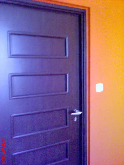 Ciemnobrązowe drzwi wewnętrzne z trzema prostokątnymi wgłębieniami na pomarańczowej ścianie, widoczny fragment klamki.