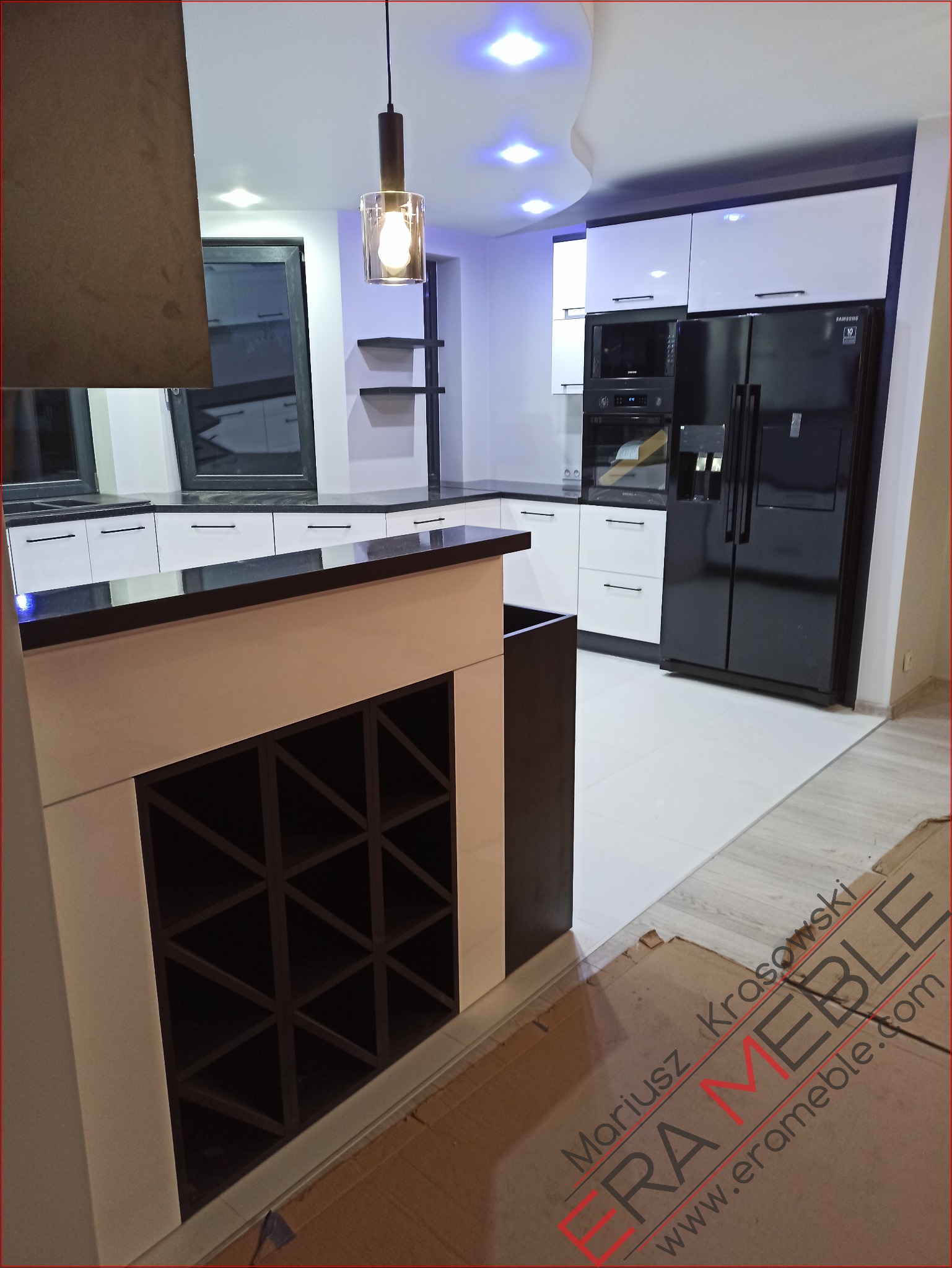 Nowoczesna kuchnia z białymi szafkami, czarnym blatem i dużą czarną lodówką, widoczny regał na wino pod blatem wyspowym, oświetlenie sufitowe punktowe i wisząca lampa z przezroczystym kloszem.