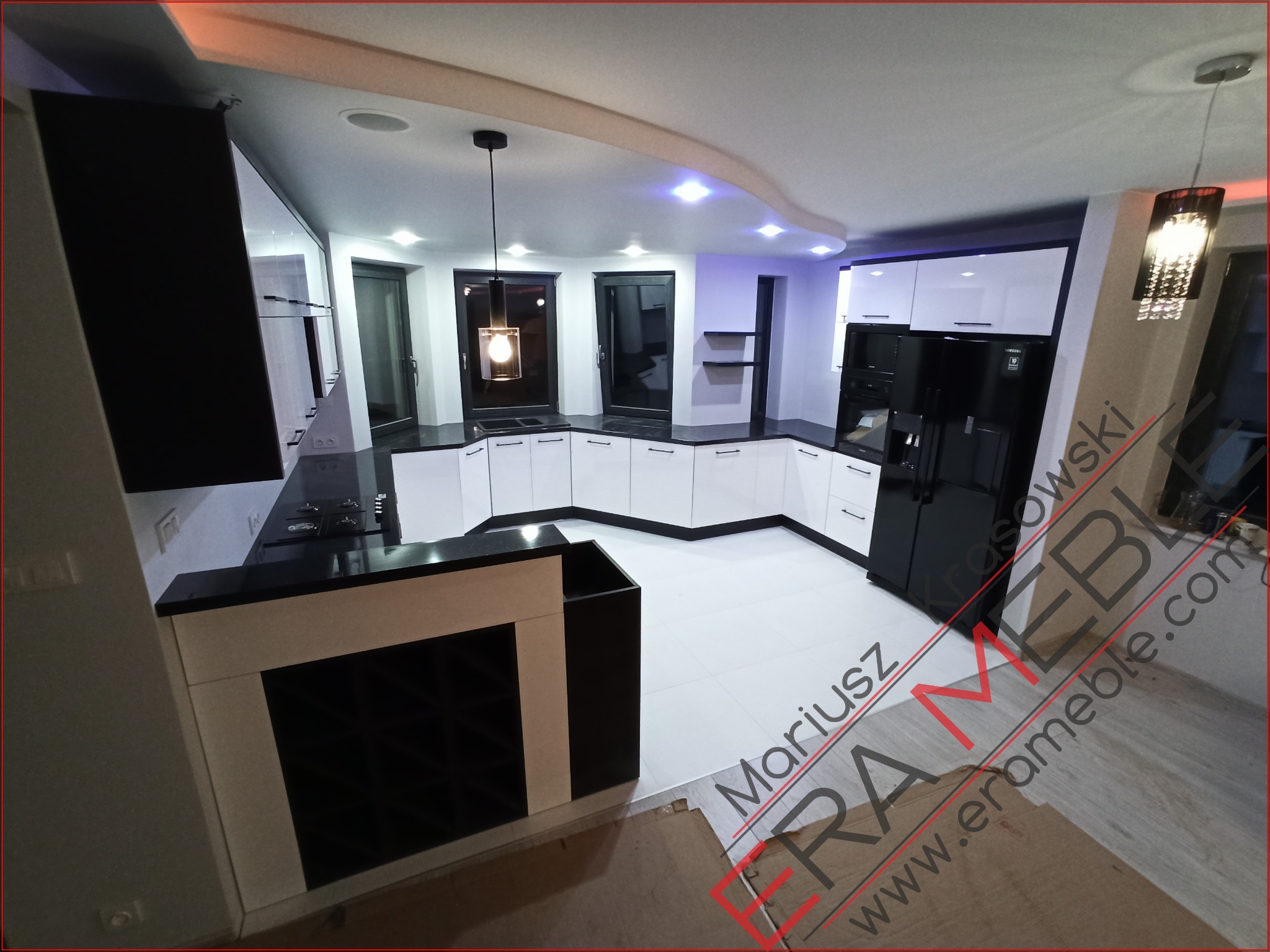 Nowoczesna kuchnia w odcieniach bieli i czerni, z czarnym blatem i białymi frontami szafek, oświetlona lampami wiszącymi i punktowymi, widok z perspektywy szerokokątnej ukazujący układ mebli...