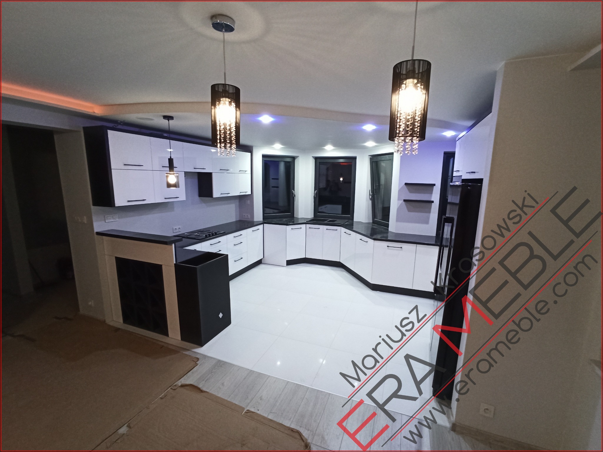 Nowoczesna, biało-czarna zabudowa kuchenna z wyspą, oświetlona wiszącymi lampami z kryształkami, widoczna z perspektywy podłogi wyłożonej białymi płytkami.