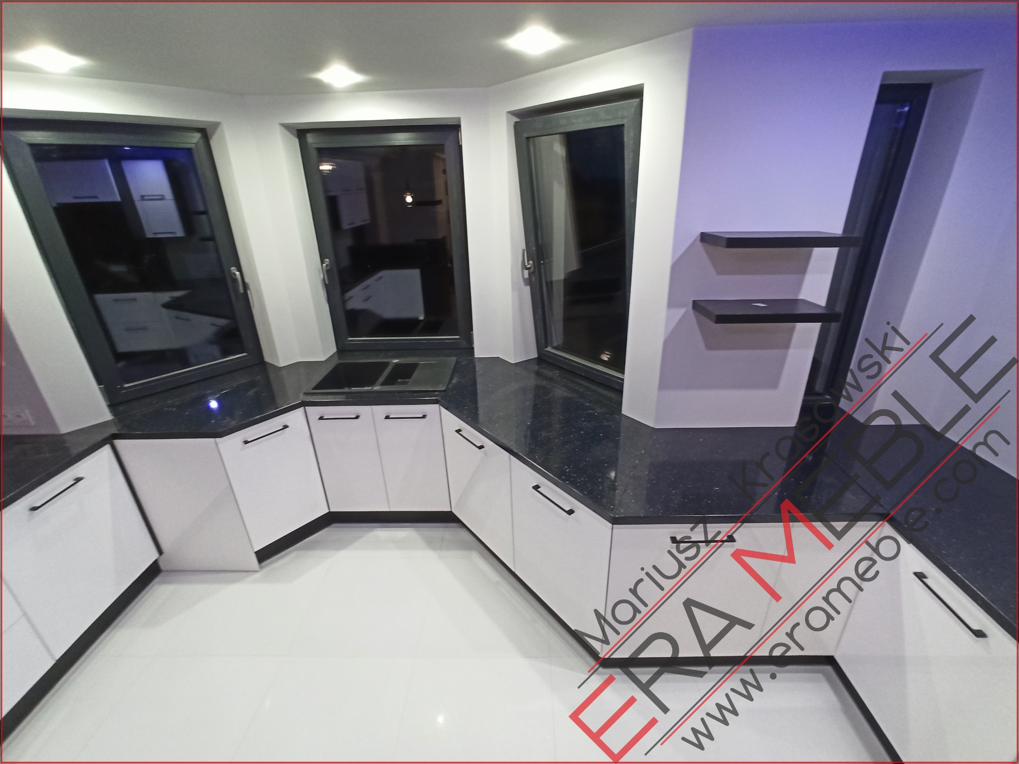 Nowoczesna kuchnia z białymi szafkami i czarnym, błyszczącym blatem, oświetlona halogenami, z trzema oknami w nieregularnym układzie i minimalistycznymi półkami na ścianie.