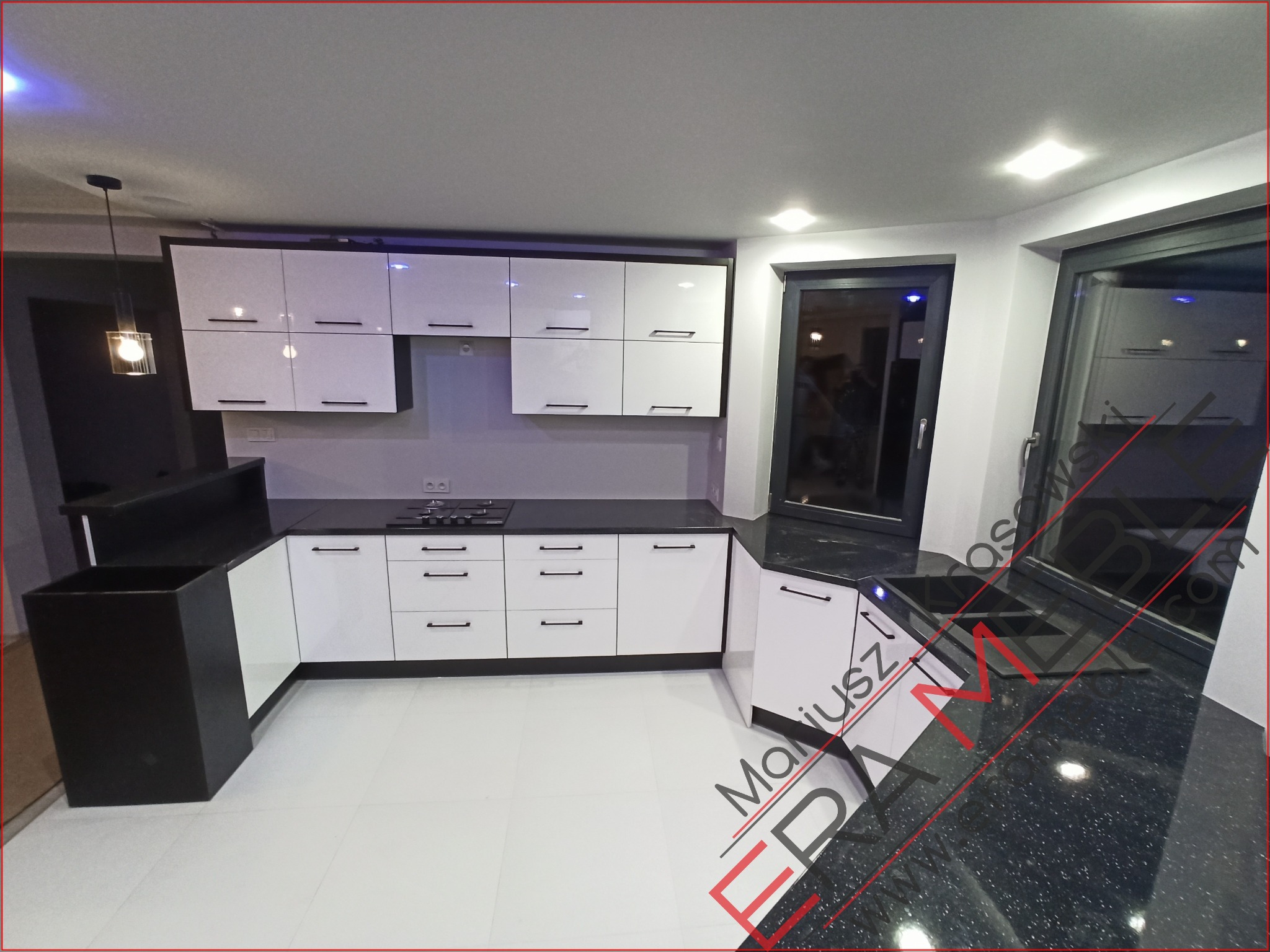 Nowoczesna kuchnia z białymi szafkami i czarnym blatem, widok na okno z czarną ramą, oświetlenie punktowe i wisząca lampa, jasna podłoga.