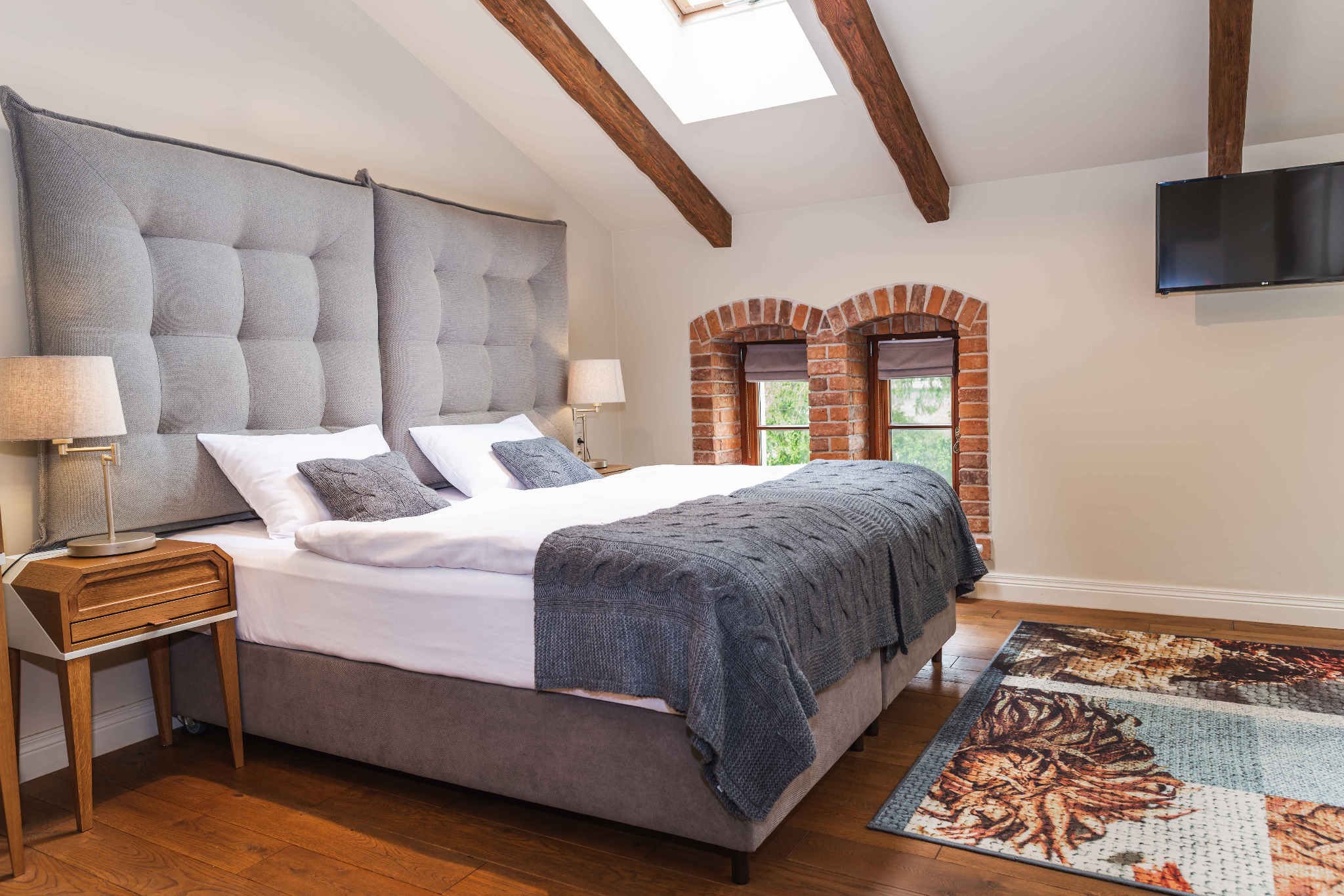 Jasna sypialnia na poddaszu z drewnianymi belkami, ceglanymi wnękami okiennymi, szarym łóżkiem z pikowanym zagłówkiem i wzorzystym dywanem.
