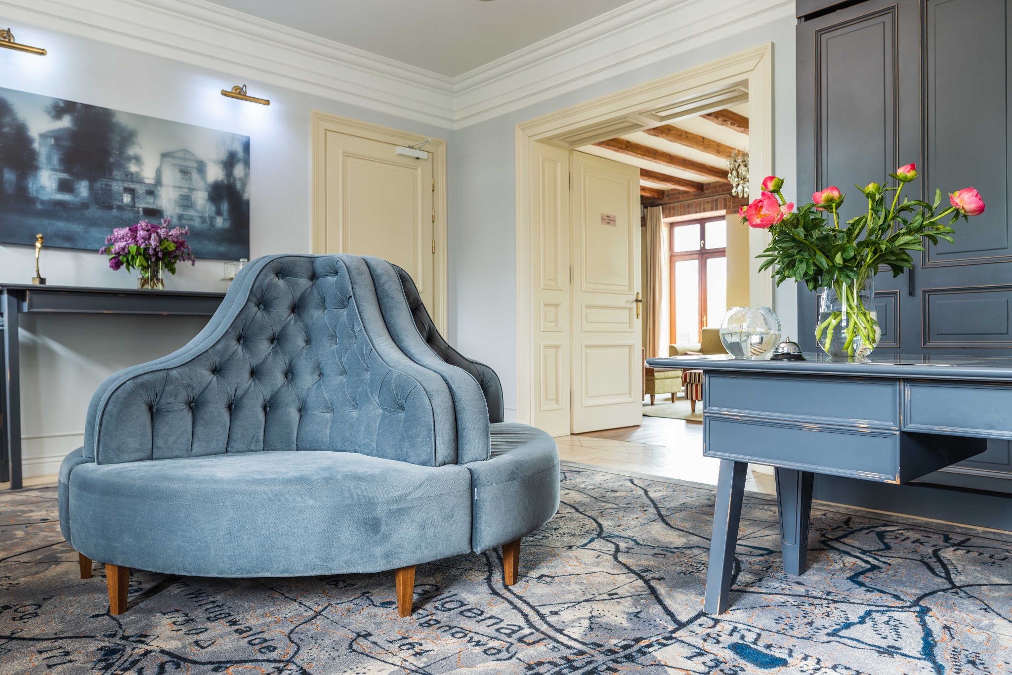 Stylowe wnętrze z okrągłą, tapicerowaną sofą w odcieniach szarości, wzorzystym dywanem z motywem mapy, ciemnoszarą konsolą z kwiatami i otwartymi drzwiami do dalszej części domu z drewnianymi...