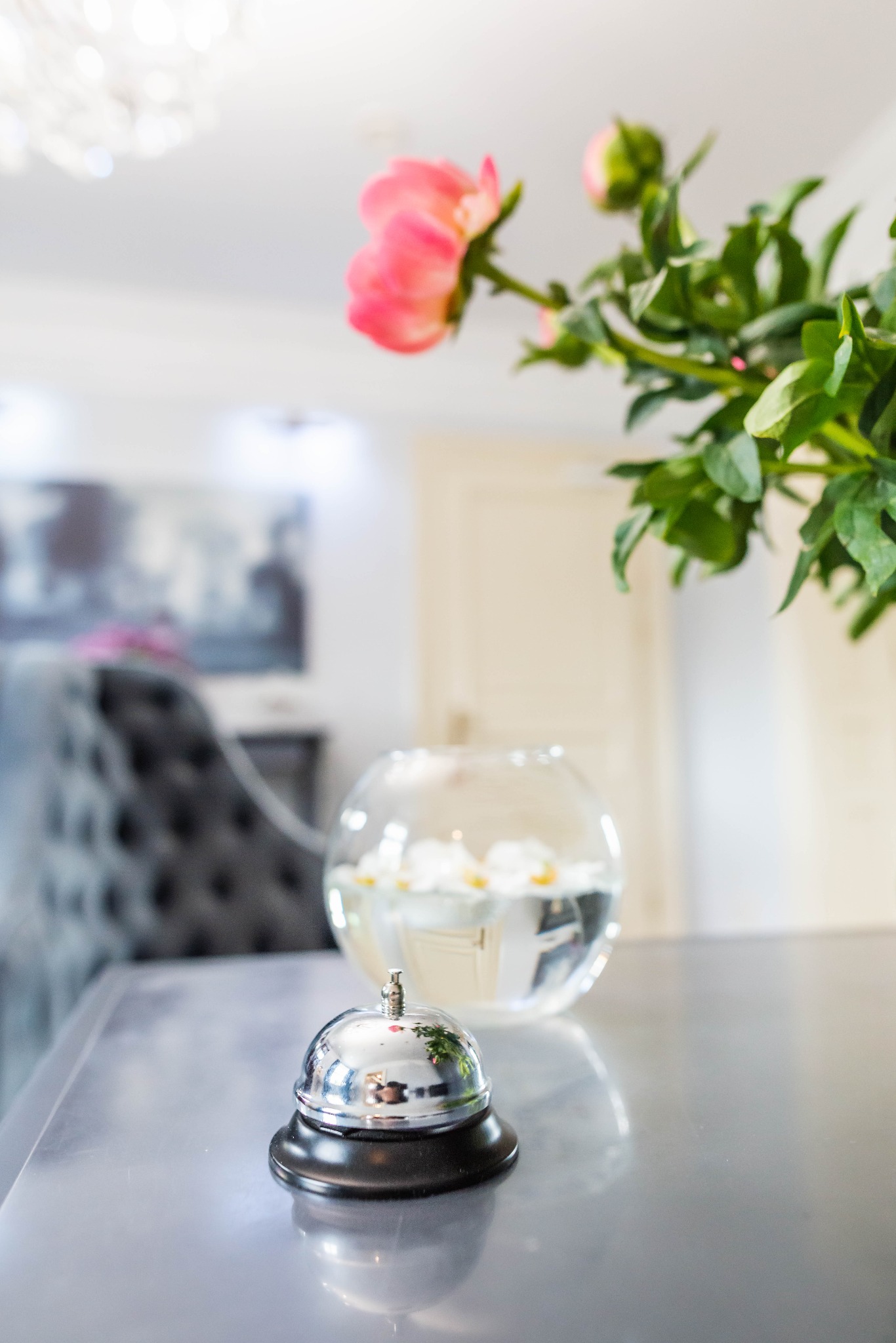 Błyszczący dzwonek hotelowy na szarym blacie stołu, w tle wazon z różowymi piwoniami i białe orchidee w szklanej kuli, za nimi szary fotel i drzwi.