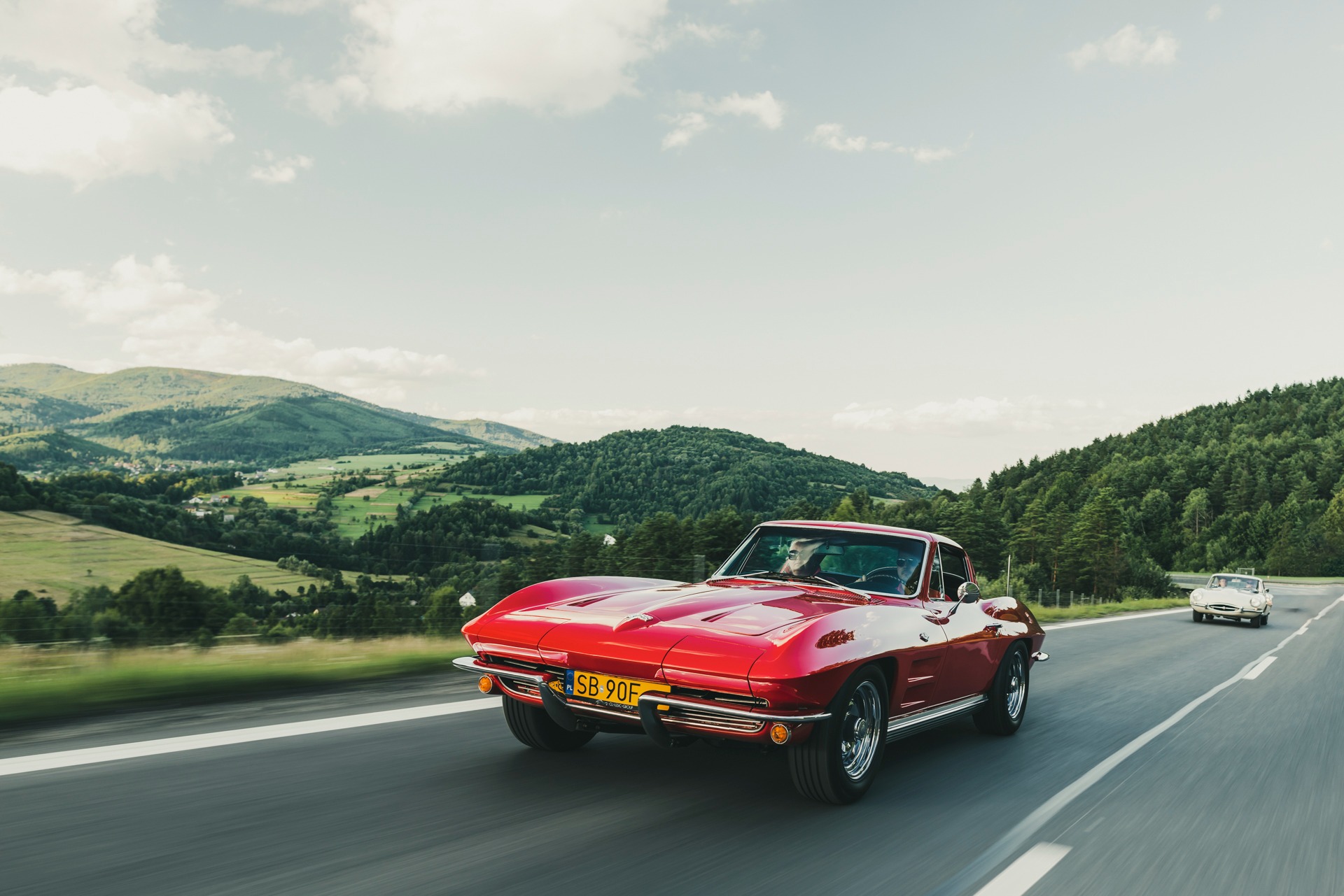 Czerwony Chevrolet Corvette C2 na drodze z widokiem na górzysty krajobraz; tablica rejestracyjna SB 90F.