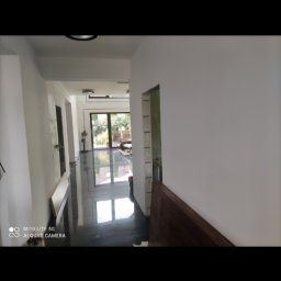 Remonty Budowlane - Korytarz w trakcie remontu, świeżo pomalowane białe ściany, czarna błyszcząca podłoga, widoczne niedokończone obramowanie drzwi i okna balkonowe z widokiem na ogród.