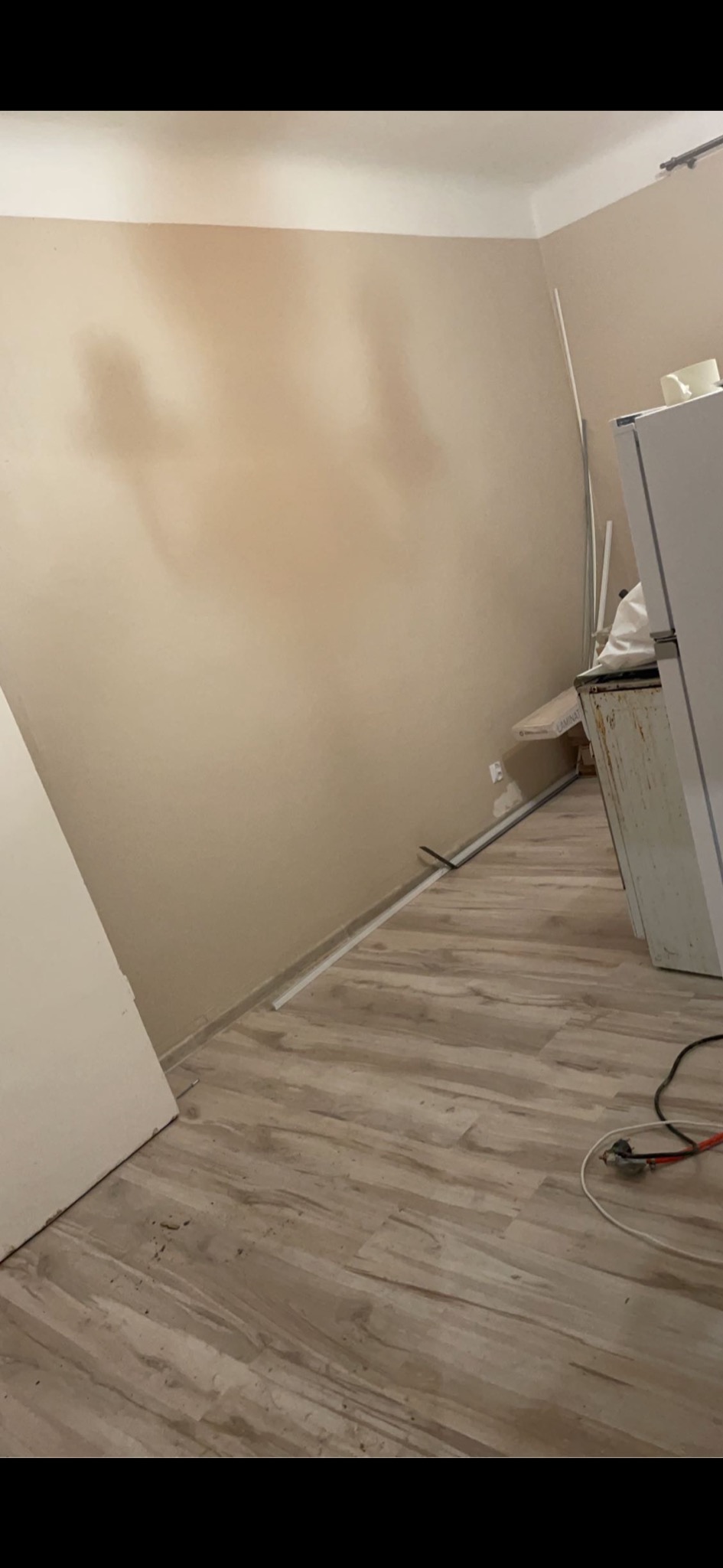 Przygotowana do malowania ściana w pomieszczeniu, z widoczną listwą przypodłogową, fragmentem drzwi i podłogą wyłożoną panelami, widoczne ślady po gładzi na ścianie.