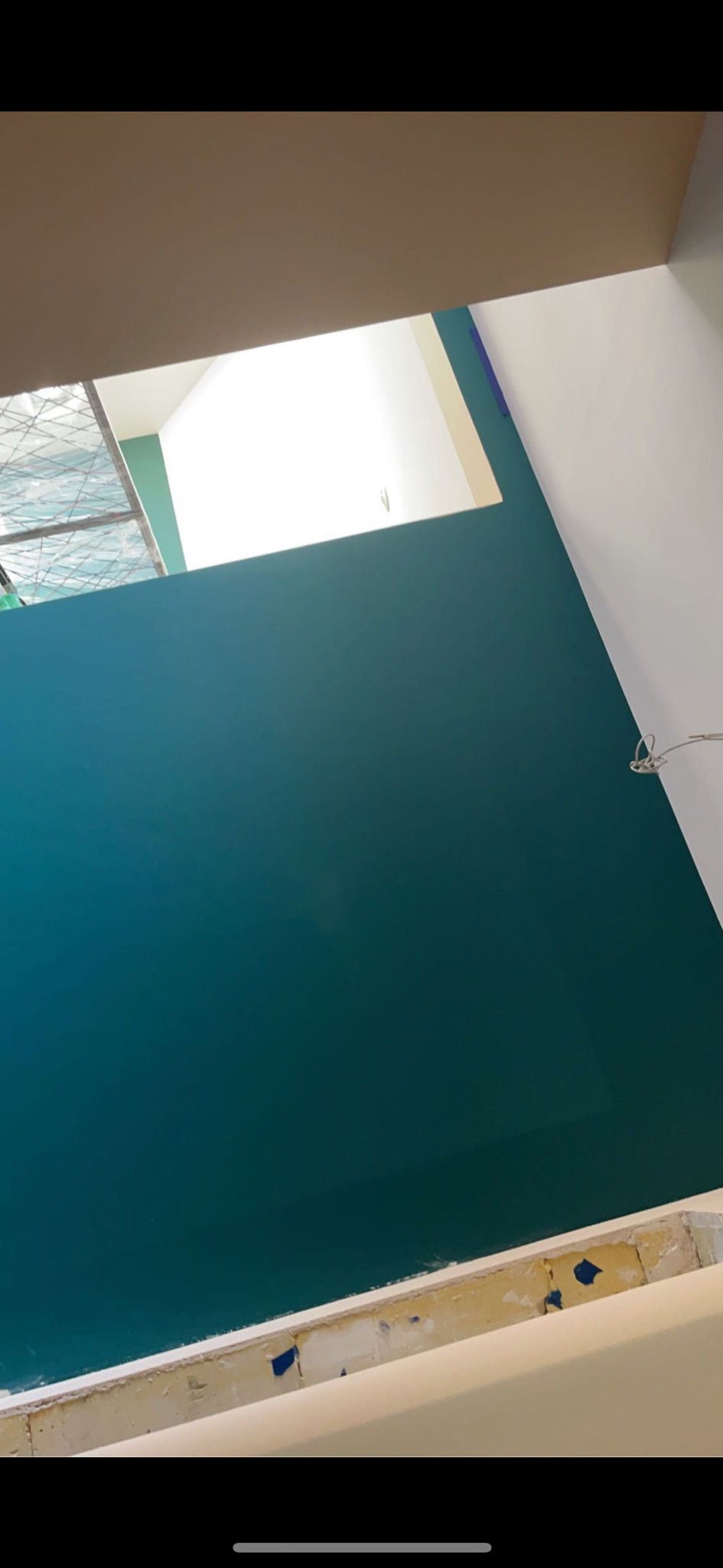 Świeżo pomalowana ściana w odcieniach turkusu, widoczna z nietypowej perspektywy, z fragmentem okna w tle i detalami wykończeniowymi w dolnej części kadru.