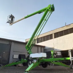 Zielony podnośnik koszowy Spierlift z operatorem w koszu na tle szarego budynku przemysłowego, drugi pracownik w kamizelce odblaskowej stoi obok.