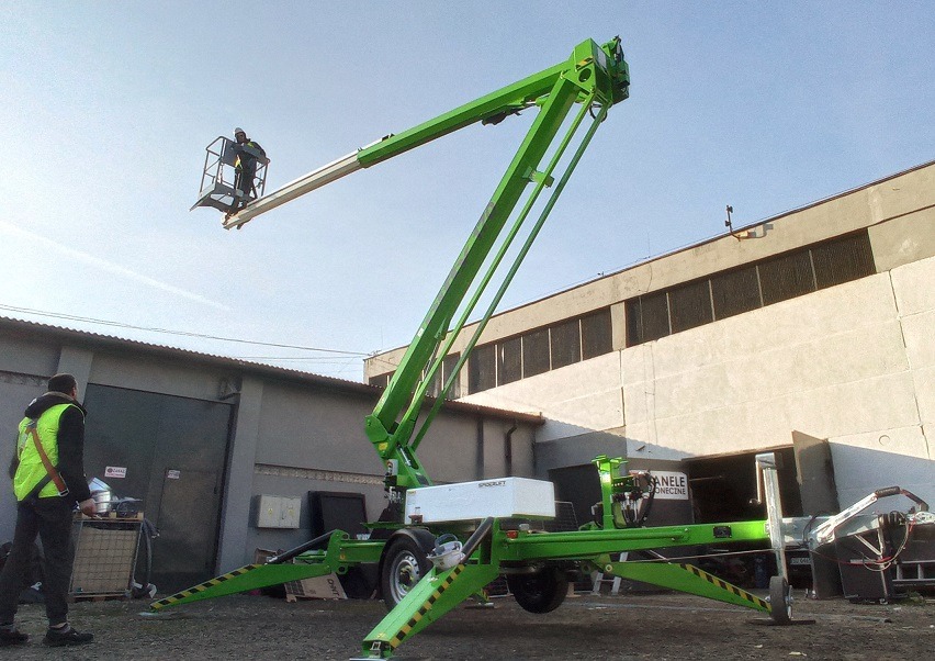 Zielony podnośnik koszowy Spierlift z operatorem w koszu na tle szarego budynku przemysłowego, drugi pracownik w kamizelce odblaskowej stoi obok.