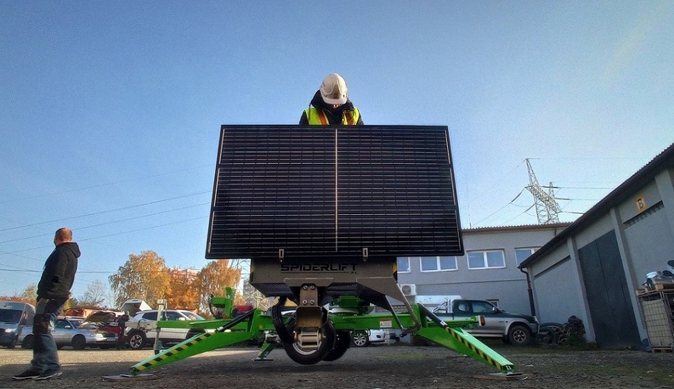 Spiderlift z panelem słonecznym w Dąbrowie Górniczej, obsługiwany przez pracownika w kasku i kamizelce odblaskowej, na tle budynku przemysłowego i pochmurnego nieba.