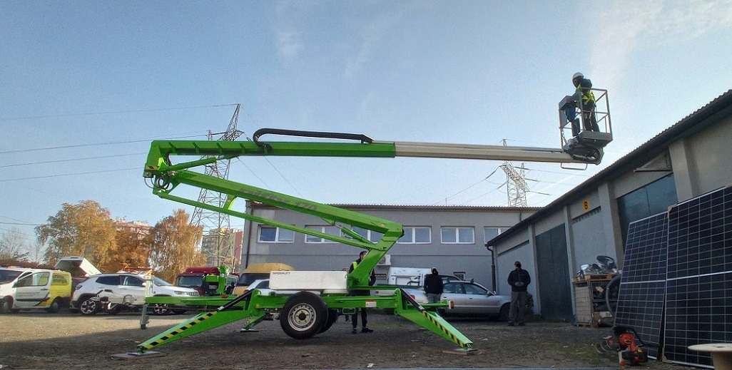 Zielona przyczepowa platforma robocza Spoolift 420 z dwoma pracownikami w koszu na wysokości, na tle budynku z panelami słonecznymi i liniami wysokiego napięcia.