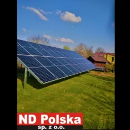 Instalacja paneli fotowoltaicznych na trawniku, widoczne podpory, w tle zabudowania i błękitne niebo z chmurami. W dolnej części logo firmy.