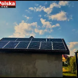 Instalacja paneli fotowoltaicznych na dachu budynku gospodarczego, widok z dołu, w tle błękitne niebo z chmurami oraz logo firmy w lewym górnym rogu.