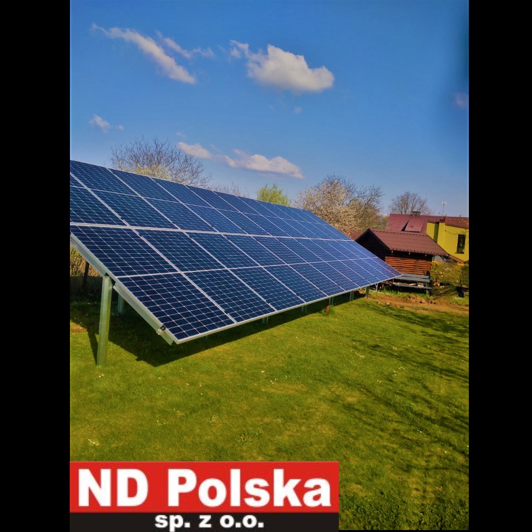 Instalacja paneli fotowoltaicznych na trawniku, widoczne podpory, w tle zabudowania i błękitne niebo z chmurami. W dolnej części logo firmy.