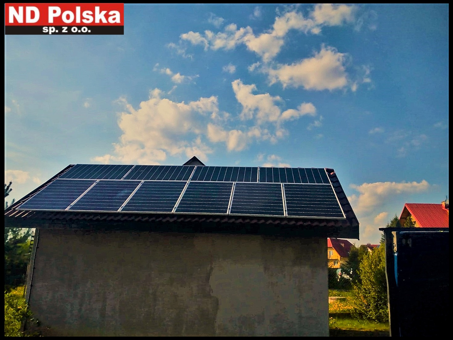 Instalacja paneli fotowoltaicznych na dachu budynku gospodarczego, widok z dołu, w tle błękitne niebo z chmurami oraz logo firmy w lewym górnym rogu.