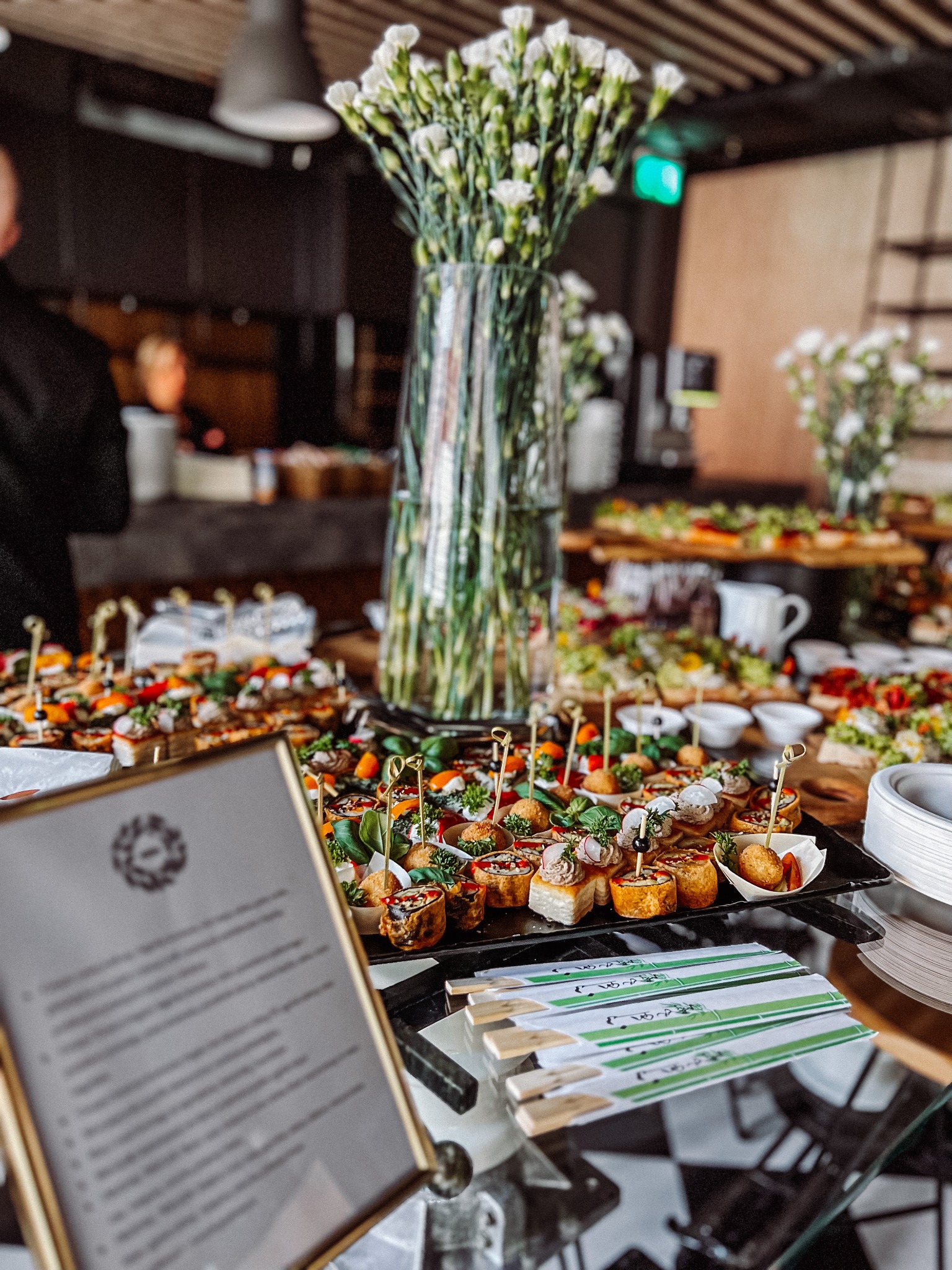 Stół z różnorodnymi przekąskami na przyjęciu, dekorowany wysokim wazonem z białymi kwiatami, widoczne menu w złotej ramce i ozdobne pałeczki.