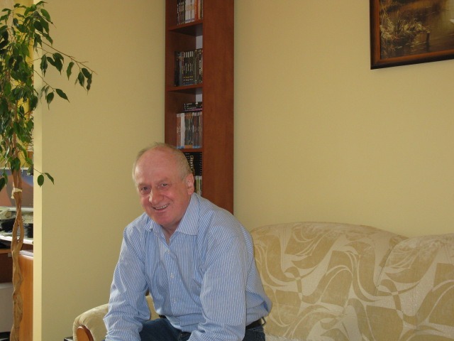 Uśmiechnięty mężczyzna w niebieskiej koszuli siedzi na kanapie w pokoju z rośliną, regałem z płytami i obrazem na ścianie.