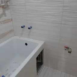 Biała wanna z zabudową w trakcie montażu, widoczne przyłącza wodne w ścianie wyłożonej płytkami z delikatnym wzorem fal, grzejnik drabinkowy w rogu.