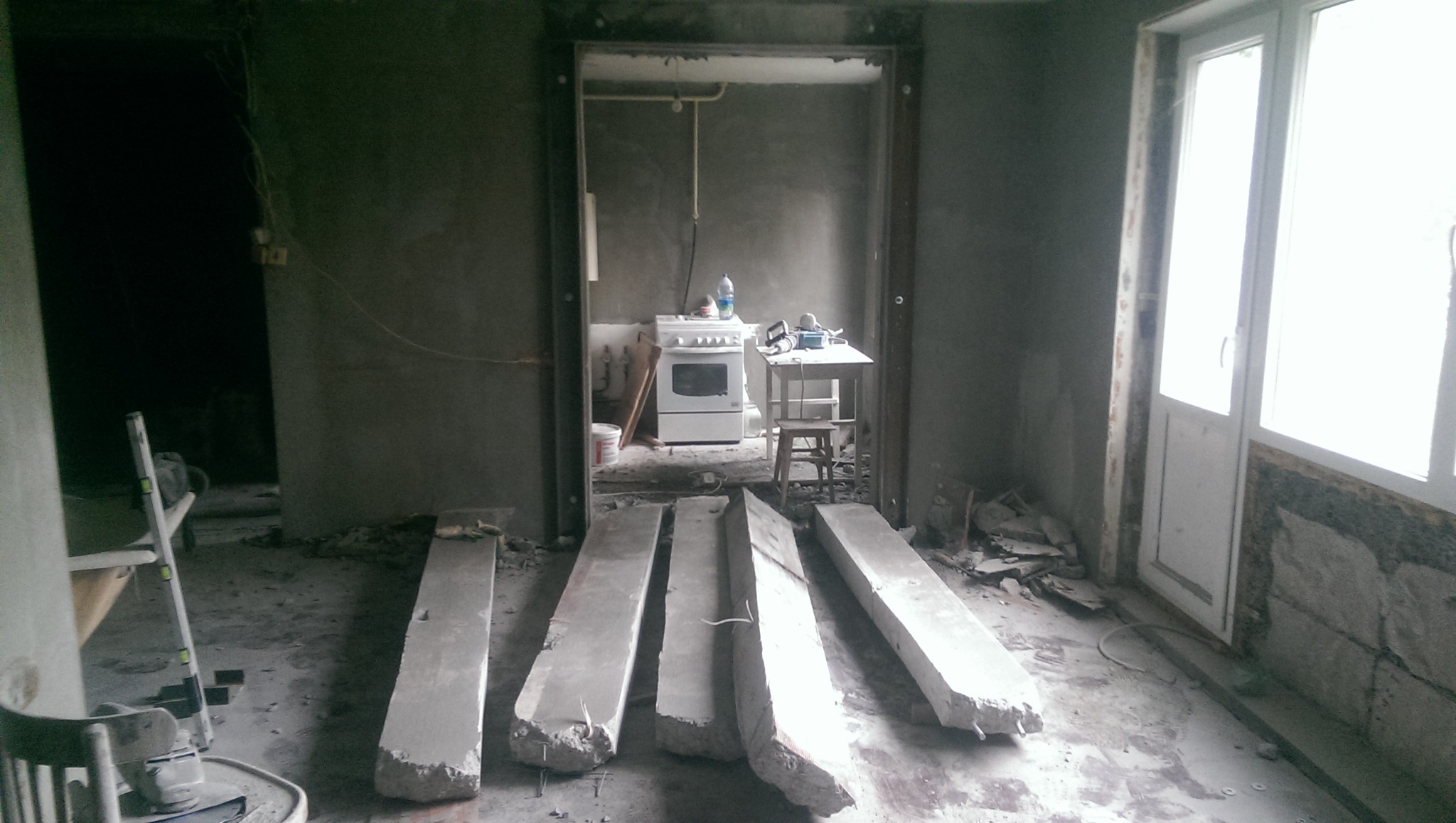 Wnętrze w trakcie remontu: fragmenty gruzu po wyburzonej ścianie leżą na podłodze, w tle widoczna kuchnia z kuchenką i stołem, surowe ściany.