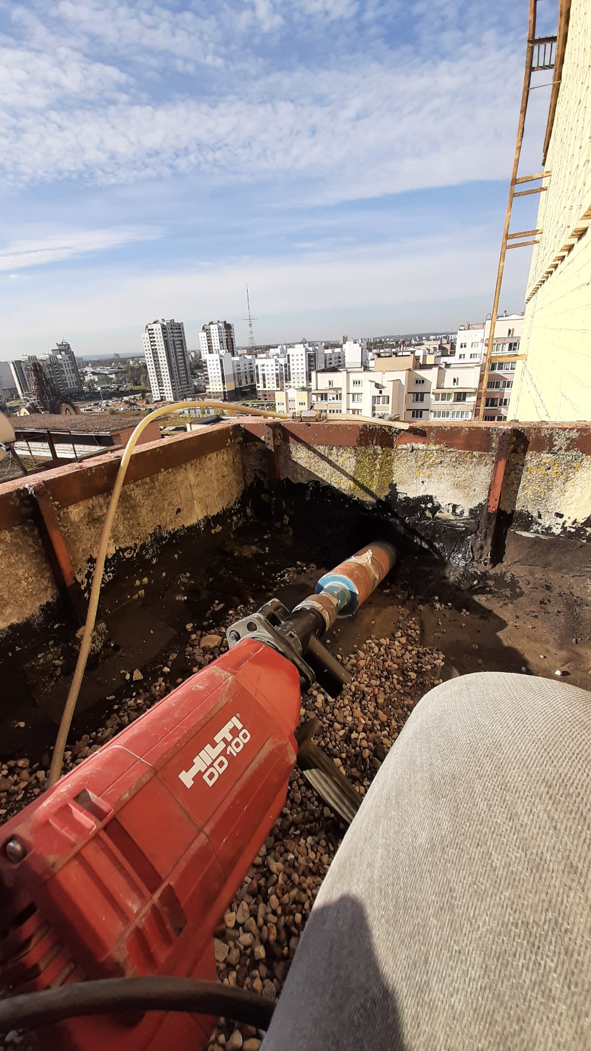 Czerwona wiertnica Hilti DD 100 wiercąca otwór w betonowej ścianie na dachu budynku z widokiem na miasto w tle.