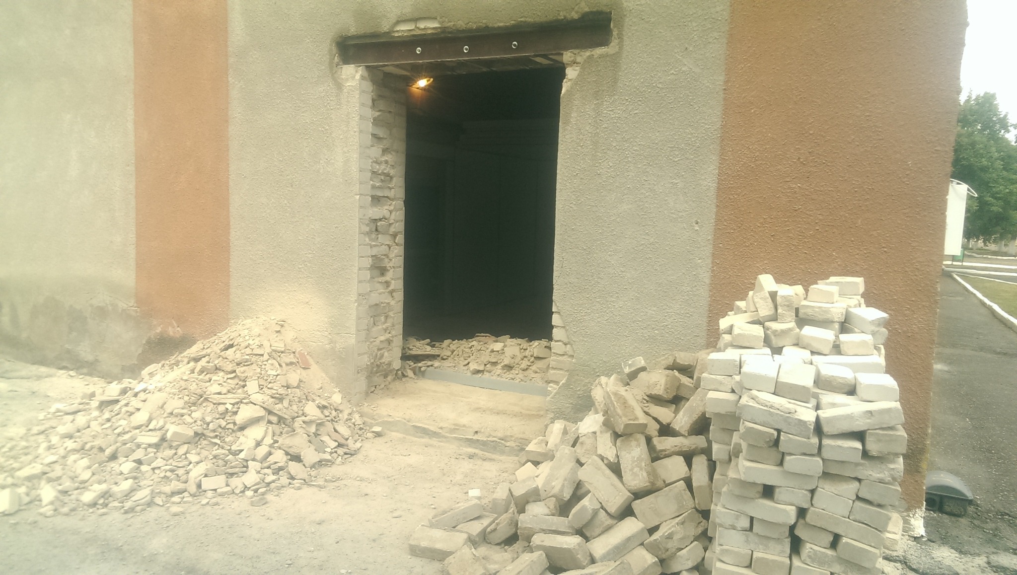 Otwór wybity w ścianie budynku z widoczną stalową belką nad nim, gruz ceglany na ziemi, w tle ciemne wnętrze pomieszczenia.