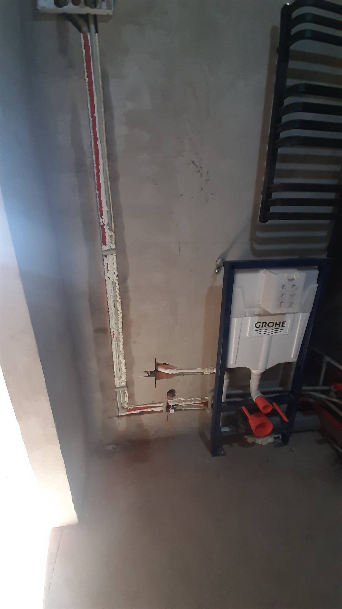 Stelaż podtynkowy Grohe zamontowany przy ścianie z widocznymi rurami instalacji wodnej zabezpieczonymi pianką montażową oraz grzejnik drabinkowy w tle.