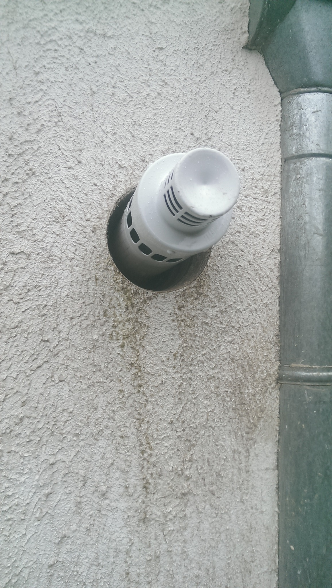 Biały, plastikowy wywiewnik kominowy wystający ze ściany o szorstkiej strukturze, obok rynna spustowa; widoczne ślady zacieków.