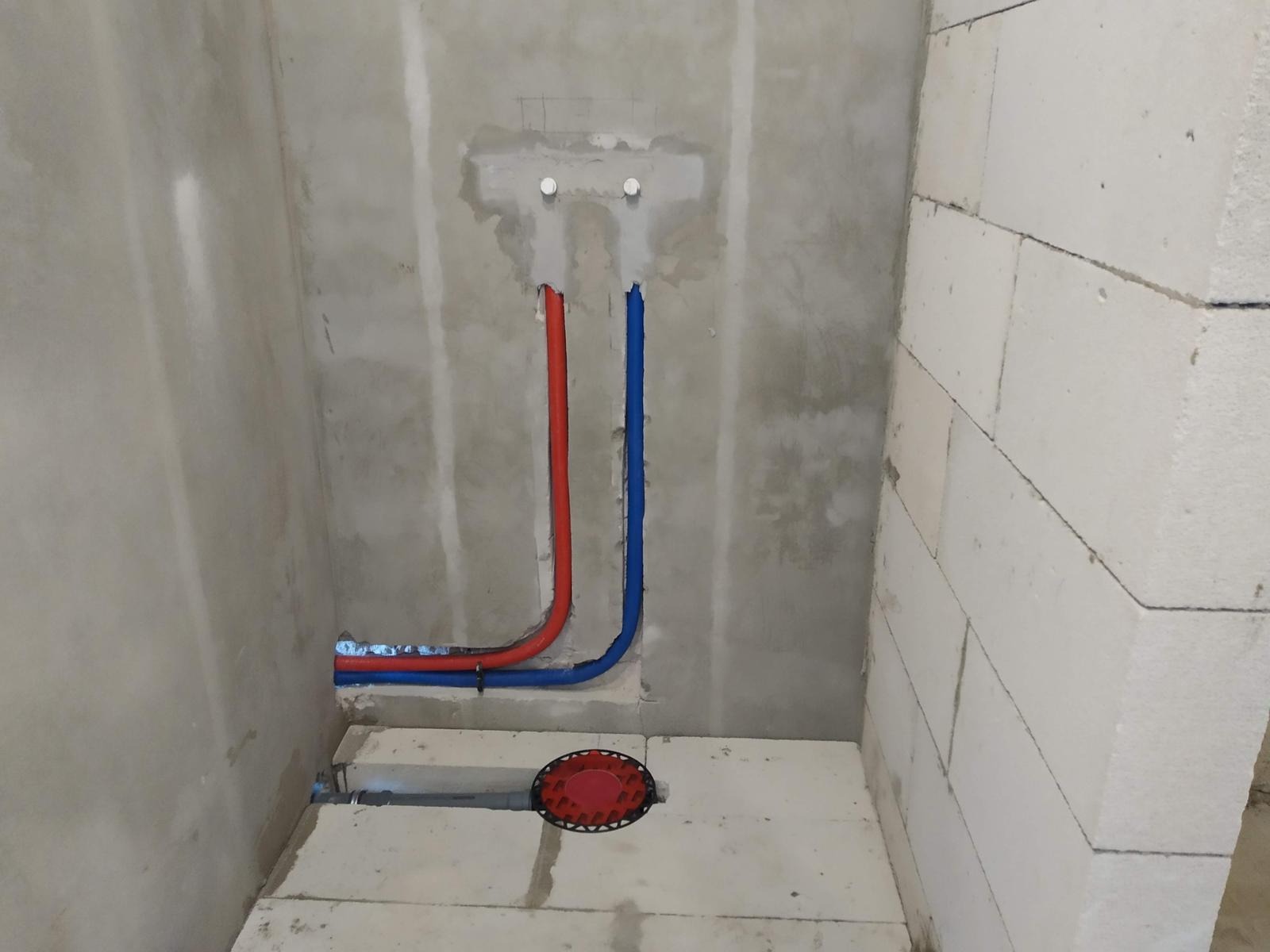 Surowy stan łazienki z widocznymi pionami instalacji wodnej (czerwony i niebieski) wychodzącymi ze ściany, oraz odpływem podłogowym z czerwonym deklem, ściany z betonu komórkowego.