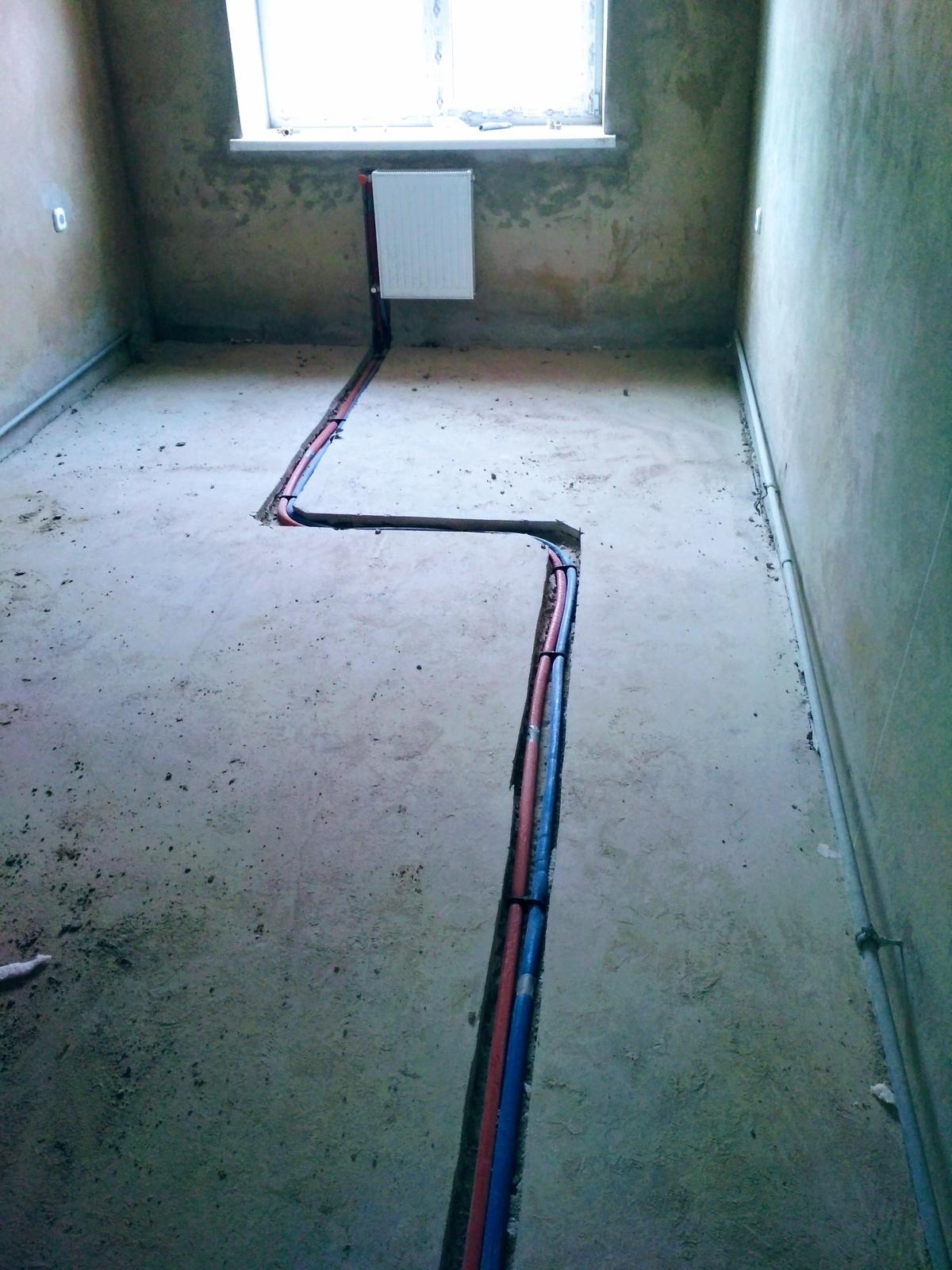 Wykute w betonowej podłodze bruzdy z ułożonymi rurami grzewczymi w kolorze niebieskim i czerwonym, prowadzące do grzejnika pod oknem w surowym wnętrzu.