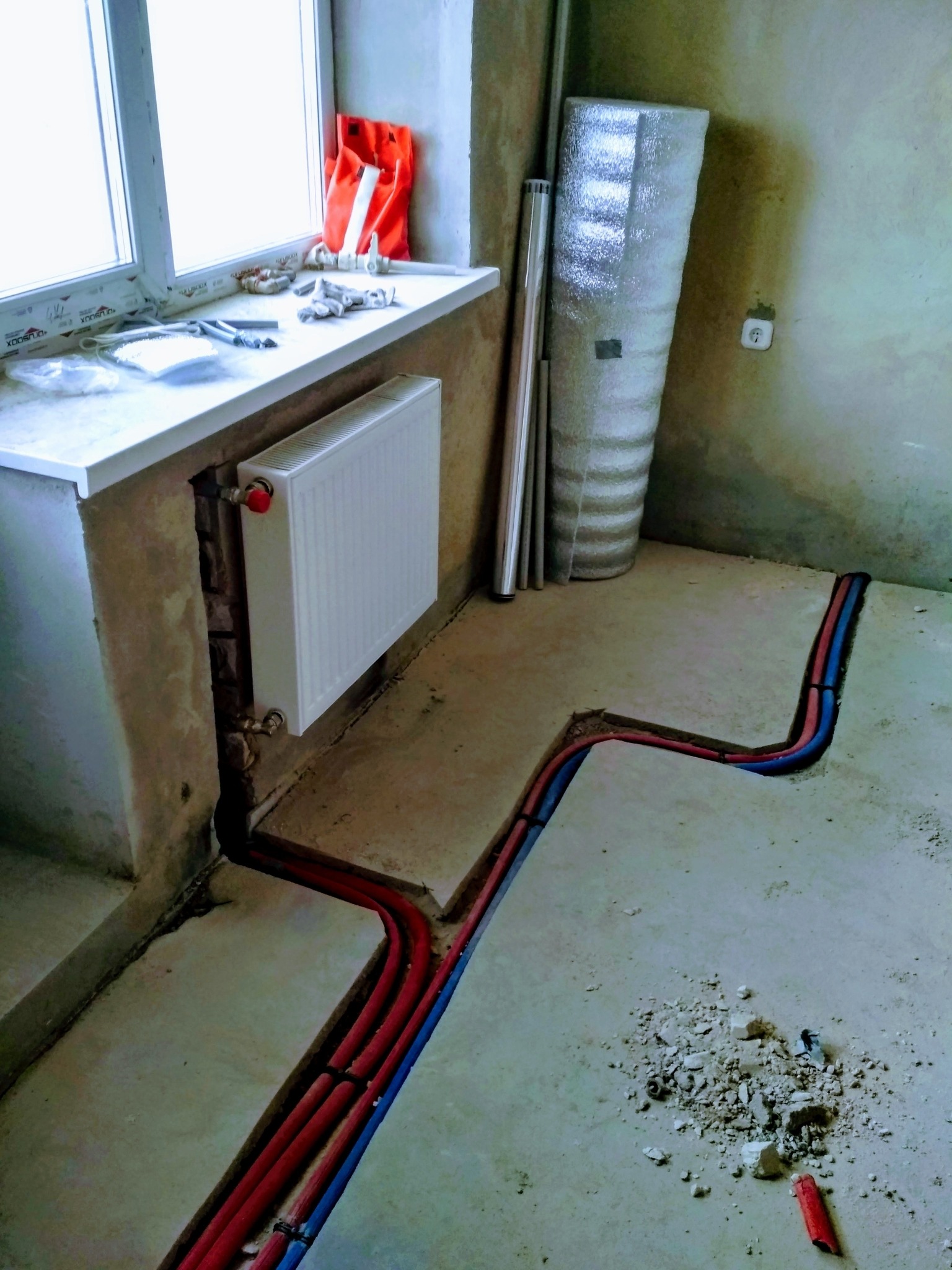 Nowy biały grzejnik zamontowany pod oknem, z widocznymi rurami grzewczymi w bruzdach w podłodze, w surowym stanie wykończenia.