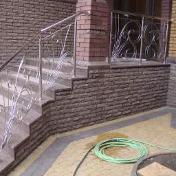 Zewnętrzne schody z balustradą ze stali nierdzewnej o ozdobnych, zakręconych elementach, prowadzące do wejścia budynku z cegły. Na pierwszym planie zwinięty wąż ogrodowy.