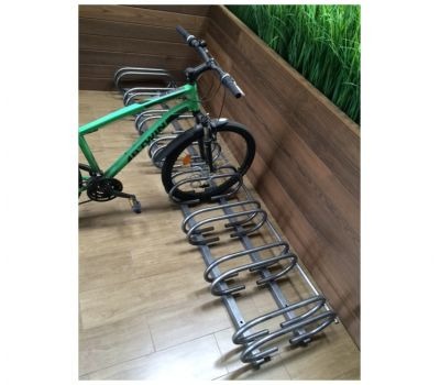 Metalowy stojak rowerowy z zaparkowanym zielonym rowerem górskim, umieszczony wewnątrz budynku z drewnianą ścianą i elementami zieleni w tle.