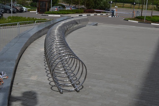 Długa, zakrzywiona, metalowa konstrukcja ze stali nierdzewnej do parkowania rowerów na zewnątrz budynku, widok z boku, z elementami miejskiej infrastruktury w tle.