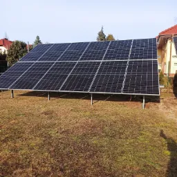 Panel fotowoltaiczny zamontowany na metalowych nogach na trawniku przed domem jednorodzinnym w słoneczny dzień.