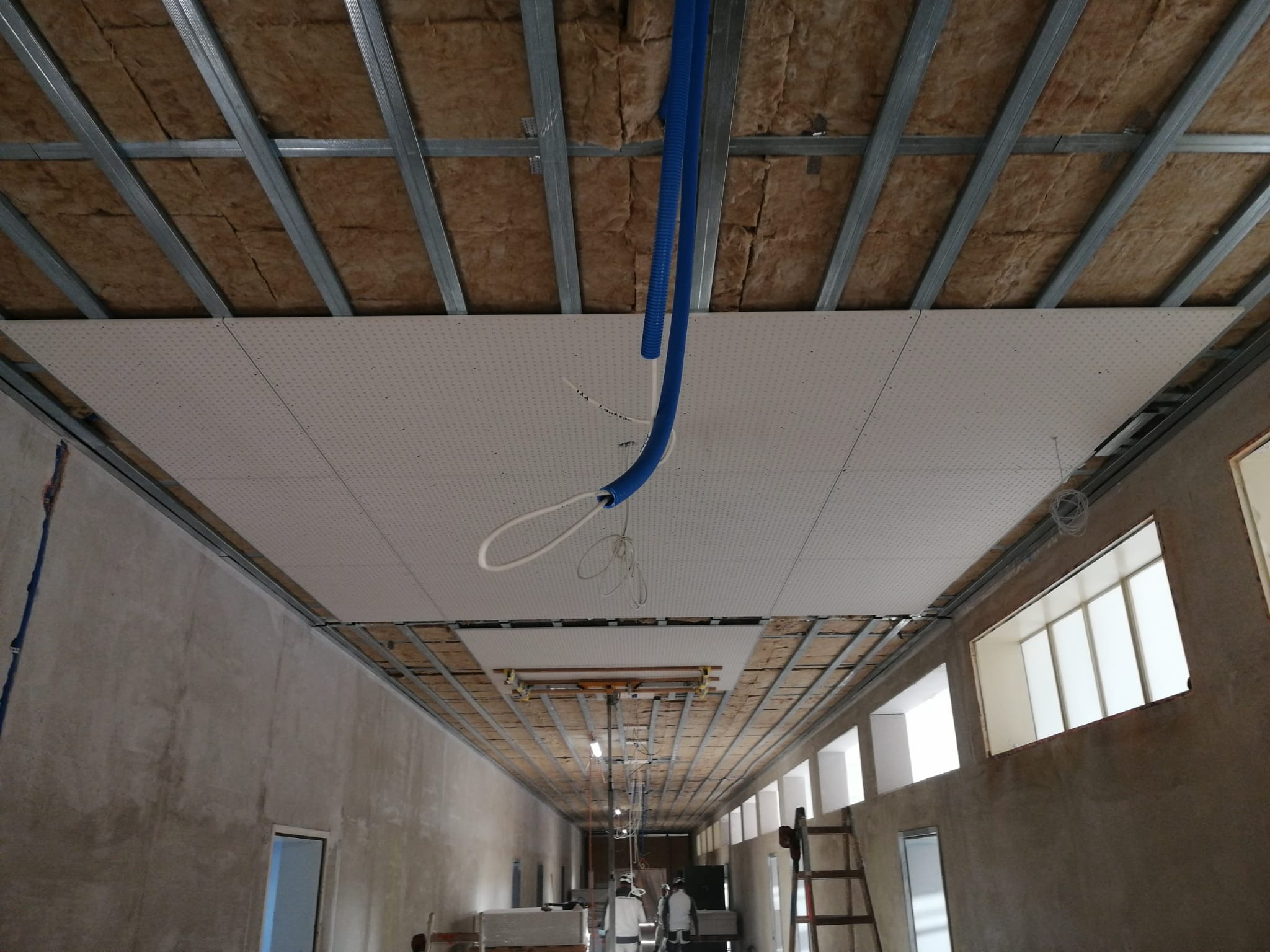 Montaż sufitu podwieszanego z widoczną konstrukcją metalową, izolacją z wełny mineralnej i częściowo zamontowanymi płytami perforowanymi, z przewodami instalacji elektrycznej i wentylacyjnej.