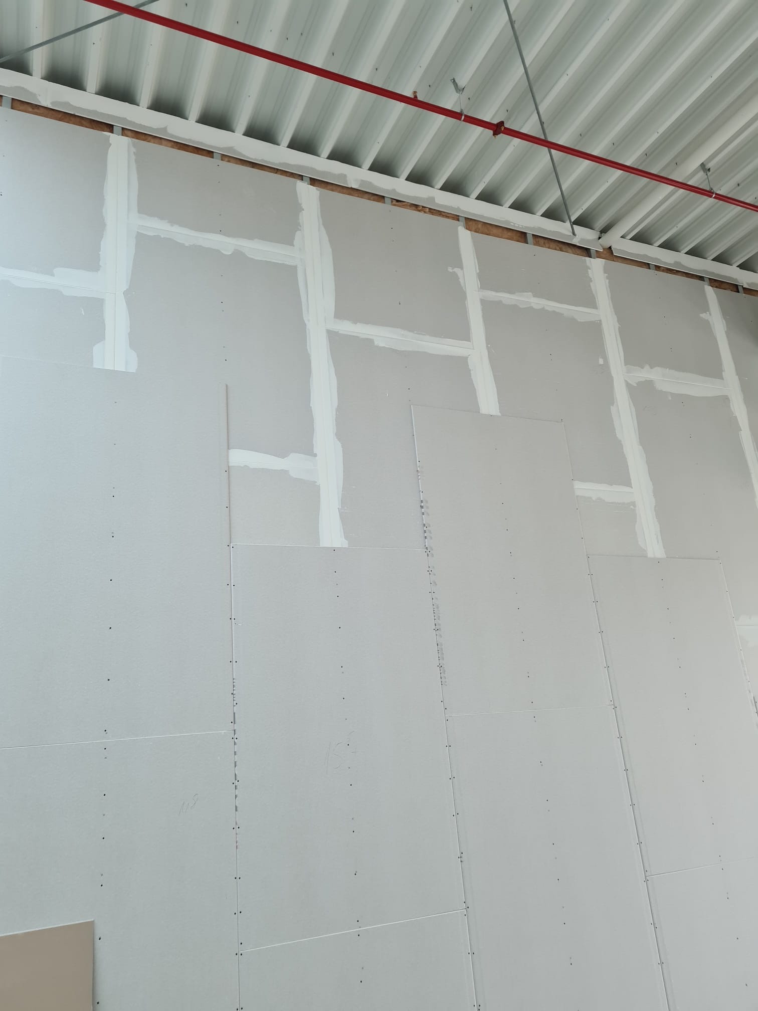 Ściana w trakcie montażu, pokryta płytami gipsowo-kartonowymi, z widocznymi łączeniami wypełnionymi masą szpachlową, oraz surowym metalowym sufitem z czerwoną rurą instalacji przeciwpożarowej.
