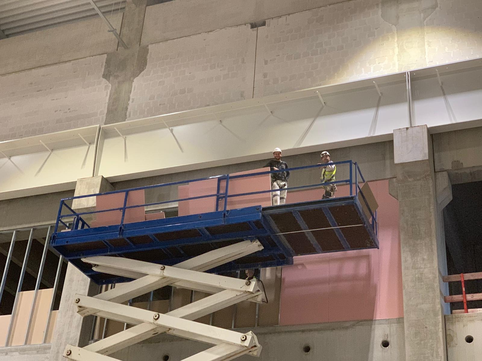 Dwóch pracowników na podnośniku koszowym montuje różowe płyty izolacyjne na ścianie w surowym wnętrzu budynku.