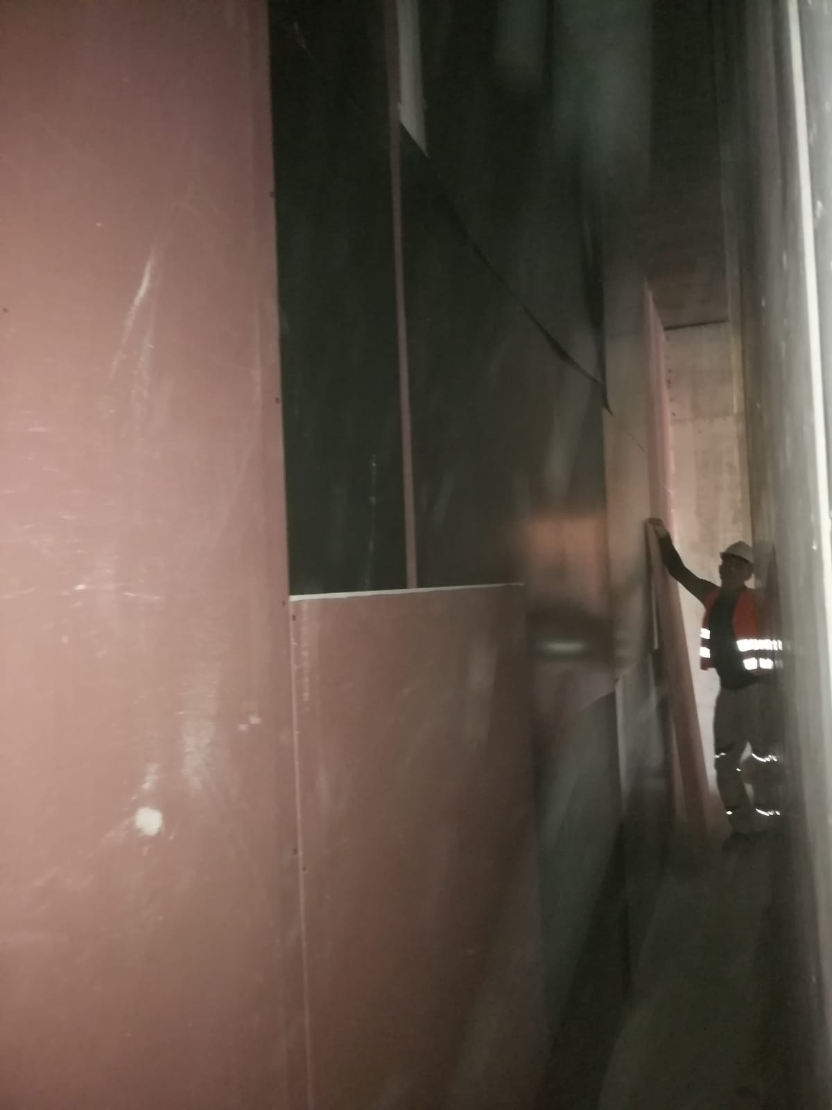 Montaż płyt karton gips w wąskim szybie, pracownik w kasku i kamizelce odblaskowej przytrzymuje płytę, widoczne ślady po szpachlowaniu na powierzchni płyty.