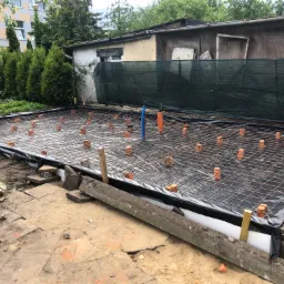 Przygotowanie zbrojenia pod płytę fundamentową, siatka zbrojeniowa na folii, podkładki z cegieł, widoczne instalacje wodne, teren budowy z elementami starej kostki brukowej i zieleni w tle.