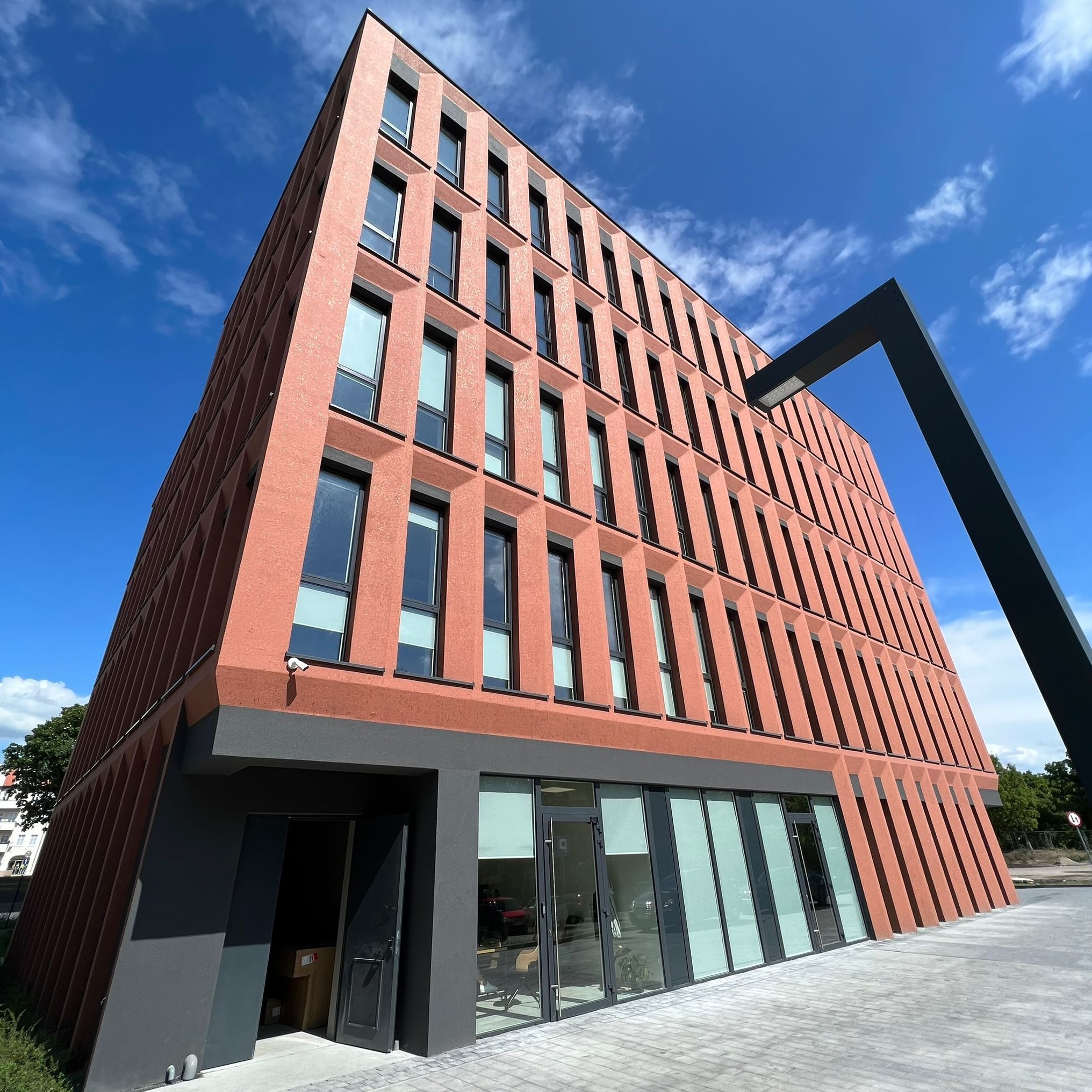 Nowoczesny budynek biurowy o geometrycznej architekturze, z ceglastą elewacją i dużymi oknami odbijającymi błękitne niebo. Ujęcie z dołu podkreśla wysokość i unikalny kształt budynku.