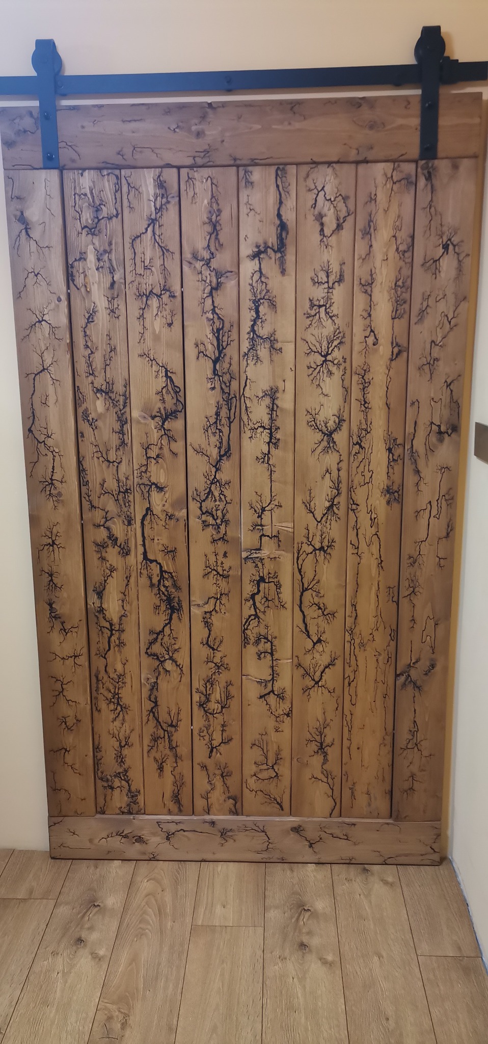 Drewniane drzwi przesuwne z unikalnym wzorem przypominającym wyładowania elektryczne, zawieszone na czarnym metalowym szynoprzewodzie.