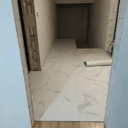 Korytarz w trakcie remontu: ułożona jasna glazura imitująca marmur na podłodze, widoczne surowe ściany z betonu komórkowego i fragmenty drewnianej podłogi, rolka zabezpieczająca na glazurze.