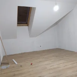 Poddasze z oknem dachowym i ułożonymi panelami podłogowymi w odcieniach drewna; widoczne listwy przypodłogowe, miernik i zabawka na podłodze, ściany pomalowane na biało.