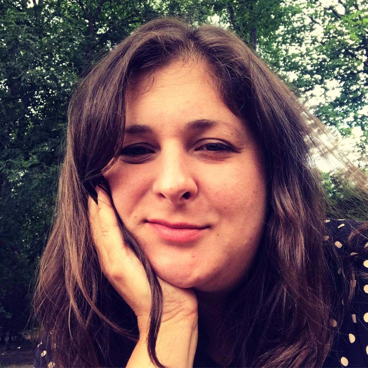 Portret uśmiechniętej kobiety z ciemnymi włosami opartymi na dłoni, w tle zielone drzewa.