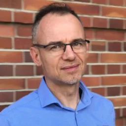 Portret mężczyzny w niebieskiej koszuli na tle ceglanej ściany. Mężczyzna ma krótkie, ciemne włosy, okulary i lekki uśmiech.