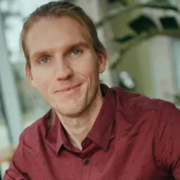 Portret uśmiechniętego mężczyzny w bordowej koszuli, z długimi blond włosami, na rozmytym tle biurowym z roślinami. Widoczny profesjonalny wyraz twarzy.