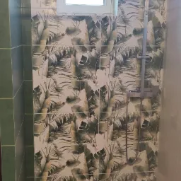 Ukończona kabina prysznicowa z deszczownicą, wyłożona płytkami z motywem tropikalnych liści, podświetlona listwą LED, z drewnopodobną podłogą i małym oknem. Widoczny fragment toalety.